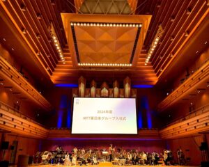 東京オペラシティコンサートホールの写真。スクリーンにNTT東日本グループ入社式と表示されている。楽団員が舞台上でリハーサルを行っている。